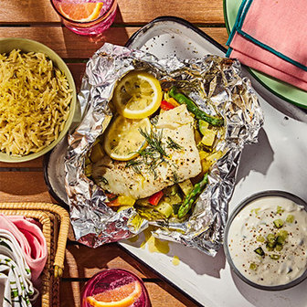 Foil packet of cooked fish with lemon slices and vegetables on a tray, served with a bowl of rice, creamy dip, and pink drinks on a wooden table.